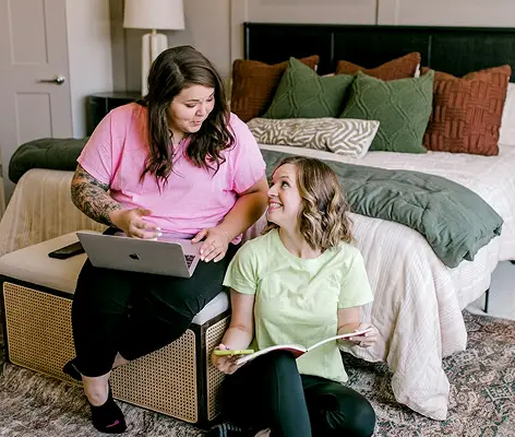Deux femmes discutant dans une chambre moderne, l'une assise avec un ordinateur portable sur les genoux et l'autre feuilletant un magazine. Elles portent des t-shirts colorés, entourées de coussins décoratifs sur le lit.