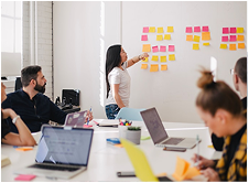 Équipe en réunion collaborative avec des post-it colorés sur un mur, une femme présente des idées tandis que d'autres travaillent sur des ordinateurs portables.