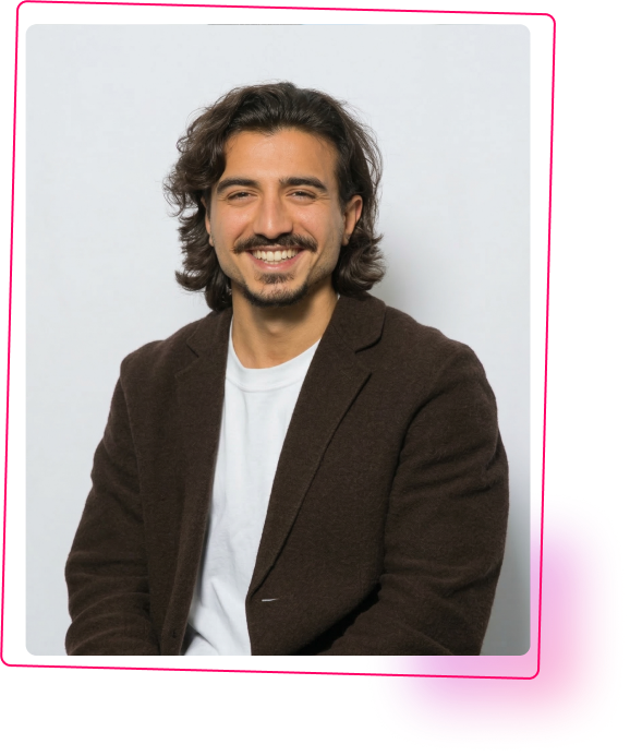 Homme souriant avec des cheveux bouclés, portant un blazer marron et un t-shirt blanc, assis devant un fond coloré.