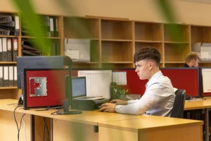 Jeune homme travaillant sur un ordinateur dans un bureau moderne, entouré de classeurs et de collègues. L'environnement de travail est bien organisé et lumineux.