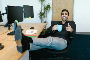 Homme souriant assis à un bureau, les pieds sur la table, tenant une tasse de café, avec deux écrans d'ordinateur en arrière-plan et une plante verte.