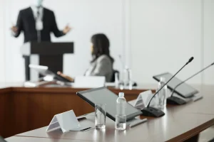 Une salle de conférence pour une formation finançable OPCO, avec des microphones, des tablettes, des bristols et des bouteilles d'eau sur la table ; à l'arrière-plan, une personne se tient debout sur un podium et une autre est assise, toutes deux hors champ.